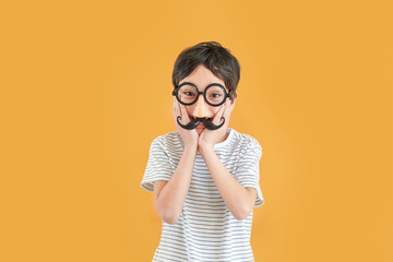 Little boy in funny disguise on color background. April fools' day celebration