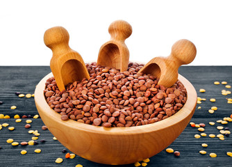 Wooden bowl of brown lentils with scoops on a wooden background.