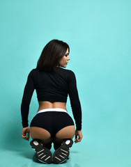 Young slim beautiful brunette woman in black sportswear and sneakers sitting back to us on floor in...
