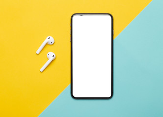 Black smartphone with blank screen and wireless earphones isolated on yellow and blue background. Clipping path.