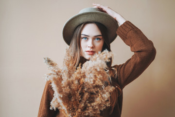 Young beautiful long brown-haired hair girl in felt hat and brown knitted sweater with dried flowers in hands on beige background