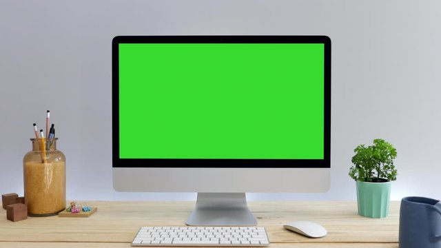 Computer desktop with mock-up green screen on wood table with grey background in office and black lamp, Zoom shoulder view.