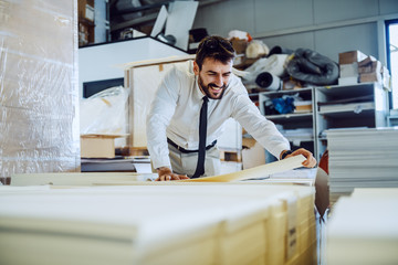 Dedicated hardworking caucasian bearded graphic engineer in shit and tie standing in warehouse and taking sheet of the pile.