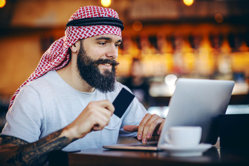 Young smiling attractive positive bearded tattooed muslim guy sitting in cafe, holding credit card and searching for something to purchase online.