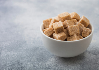 White bowl of natural brown sugar cubes on light table background.