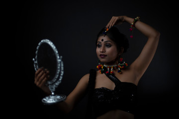Fashion portrait of Indian Bengali brunette woman in Indian traditional tribal dress and handmade ornaments doing makeup in dark copy space studio background. Indian lifestyle and fashion photography.