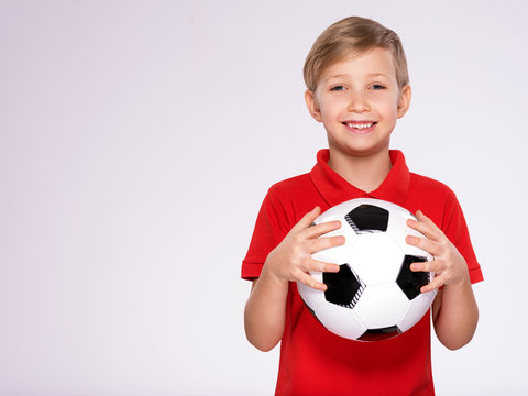 Photo Of A Smiling Boy In Sportswear Holding Soccer Ball, Posing At Studio. Happy 8 Years Old Kid In A Red T-shirt With A Soccer Ball In Hand. White Child With A Smile Holds A Soccer Ball.