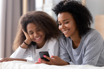 Happy african mother and daughter using smartphone lying on bed