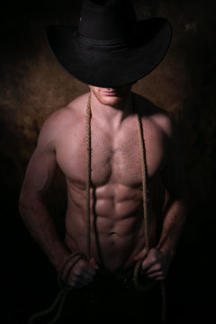 Shirtless cowboy wearing hat covering his face, holding rope around his neck with defined pecs and muscular sixpack abs