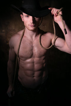 Handsome cowboy wearing a hat with barechest showing defined pecs and sixpack abs, muscular arms holding onto rope