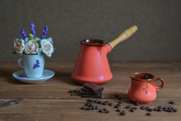 Ceramic cup, cezve, coffee grains, wooden background and pieces of chocolate, flower on the background.