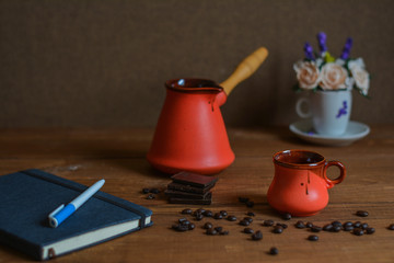 Ceramic cup, cezve, coffee grains, wooden background and pieces of chocolate, notebook with pen, flower.