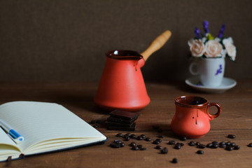 Ceramic cup, cezve, coffee grains, wooden background and pieces of chocolate, notebook with pen, flower.