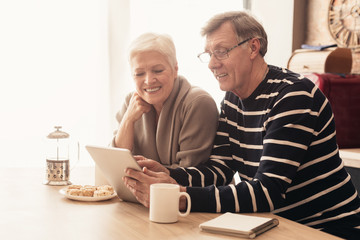 Cheerful elderly couple using digital tablet in kitchen together, reading news