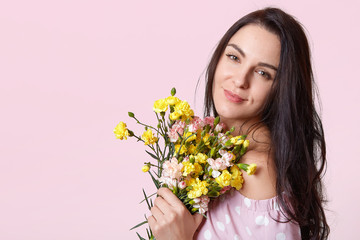 Obraz na płótnie Canvas Indoor shot of young adorable brunette woman with bouquet of yellow flowers, gentle girl wearing pink sundresses with bare shoulders, model looking directly at camera, isolated over rosy wall.