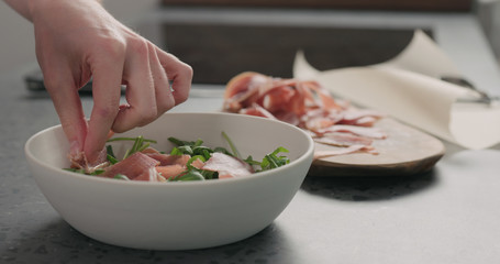 man hand adding prosciutto to make salad with arugula, and tomatoes