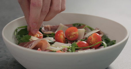 man add fresh cherry tomatoes in white bowl with mixed salad leaves and prosciutto