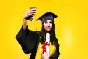 graduate woman makes selfie photo on phone over yellow background, concept of successful completion...