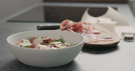 pour olive oil over salad with prosciutto, cherry tomatoes and mixed salad leaves in white bowl