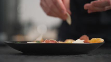 making salad man adding parmesan into black plate with arugula, prosciutto and tangerine