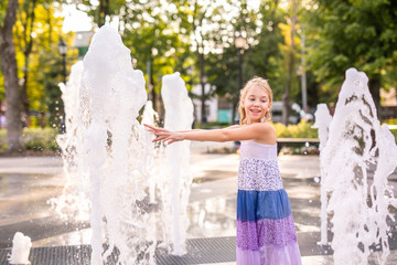 Blonde happy child girl in long purple dress running between water flow in city summer park. Child activity concept