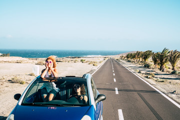 Travel friends and transport with blue convertible car and couple of adult women friends have fun together driving on a long road with ocean in background - youth female stand up outside the roof