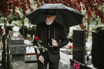 Elegant sad elderly man standing on the rain with umbrella and grieves at the grave of a loved person