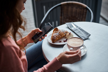 Woman have breakfast in caffe and using smartphone. Girl chatting and using internet with phone during coffee break with a croissant