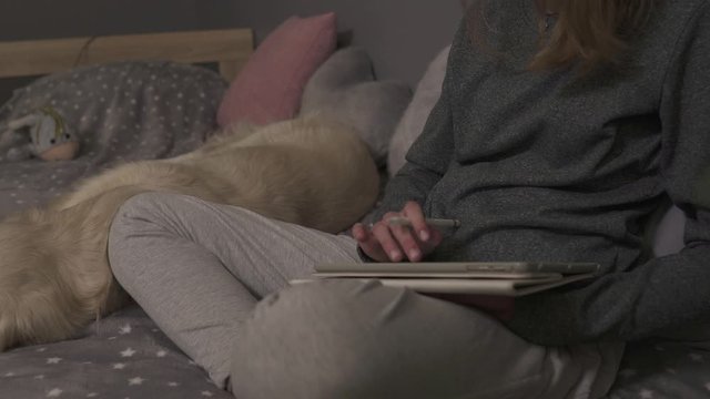 Girl sitting on a bed in an room drawing with her dog.