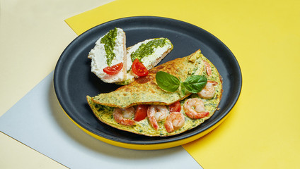 Continental breakfast - omelet with shrimps, tomatoes and basil with toast with cream cheese and pesto on a black plate on colored backgrounds.