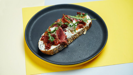 Trendy street snack. Tasty sandwich with jamon, poached eggs, cucumber and microgreen on black plate on colored background. copy space.