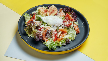 Selective focus on served Caesar salad with Salmon in black plate on colored background. Tasty seafood salad