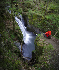 Young man in orange jacket looking down on waterfall near Ulmen in Eifel county, Germany