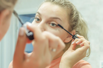 the blonde girl paints her eyelashes with mascara at home in front of the mirror. close up. Selective focus, film grain.