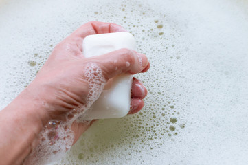 A piece of white soap in the hand of a man on a background of soap suds. Concept of hygiene and body care.