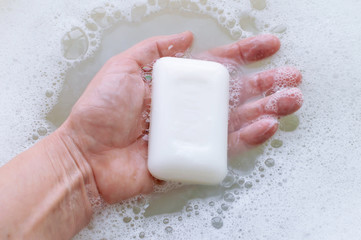 A piece of white soap in the hand of a man on a background of soap suds. Concept of hygiene and body care.
