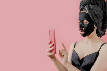 Young woman apply black cosmetic fabric facial mask and phone in hands on pink background. Face peeling mask with charcoal, spa beauty treatment, skincare, cosmetology. Close up