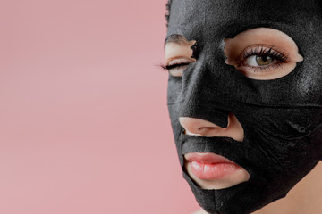 Young woman apply black cosmetic fabric facial mask on pink background. Face peeling mask with charcoal, spa beauty treatment, skincare, cosmetology. Close up