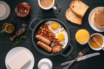 English breakfast on a dark table. fried eggs with bacon canned beans and sausage. near coffee, orange juice and pancakes with butter and honey