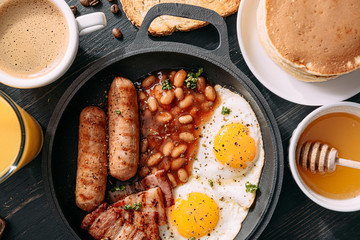 English breakfast on a dark table. fried eggs with bacon canned beans and sausage. near coffee, orange juice and pancakes with butter and honey