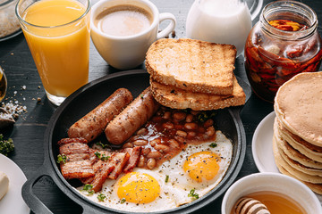 English breakfast on a dark table. fried eggs with bacon canned beans and sausage. near coffee, orange juice and pancakes with butter and honey