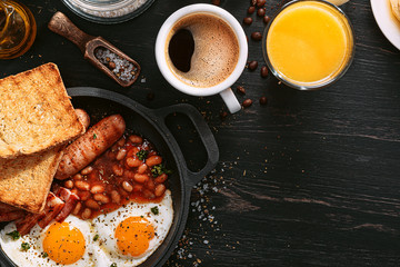 English breakfast on a dark table. fried eggs with bacon canned beans and sausage. near coffee, orange juice and pancakes with butter and honey
