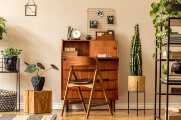 Retro interior design of living room with wooden vintage bureau, shelf, cube, plants, cacti, books, decoration and elegant personal accessories. Stylish vintage home decor. Beige wall. Template. 