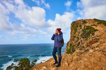 Young woman tourist at Cape Cabo da Roca standing on the rocky edge of the cliff. The waves of the ocean break on the rocks at the bottom of the cliff