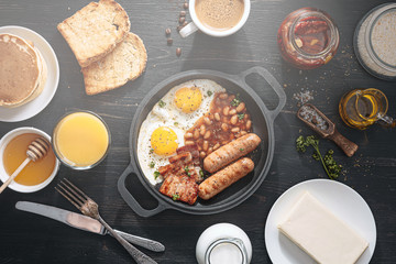 English breakfast on a dark table. fried eggs with bacon canned beans and sausage. near coffee, orange juice and pancakes with butter and honey