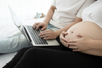 Naklejka premium Young man and his pregnant sat on the bed while using a notebook computer to find information about childbirth and preparation before going to consulting doctor the hospital. Technology concept.