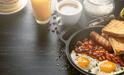 English breakfast on a dark table. fried eggs with bacon canned beans and sausage. near coffee, orange juice and pancakes with butter and honey