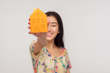 Property purchase, reliable real estate agency. Happy cheerful asian girl with brunette hair in summer blouse showing paper house in outstretched hand and smiling. indoor studio shot, pink background