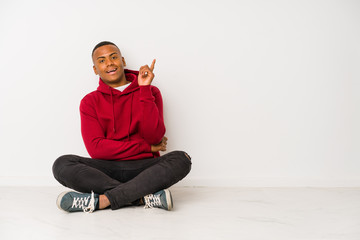 Young latin man sitting on the floor isolated smiling cheerfully pointing with forefinger away.