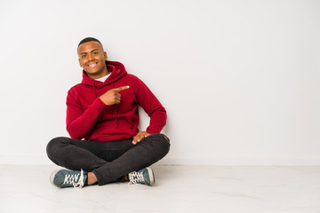 Young latin man sitting on the floor isolated smiling and pointing aside, showing something at blank space.